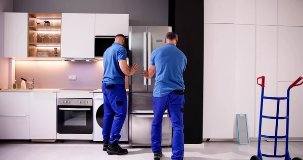Deux techniciens en uniforme bleu installent un réfrigérateur américain inox dans une cuisine moderne blanche